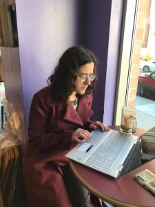 Picture of author writing on a laptop, in a coffee shop, wearing a long red coat. There is a coffee beside her on the table.