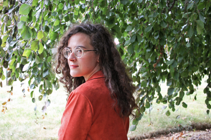 Author sits beneath a leafy tree, smiling, in a rust-orange blazer.