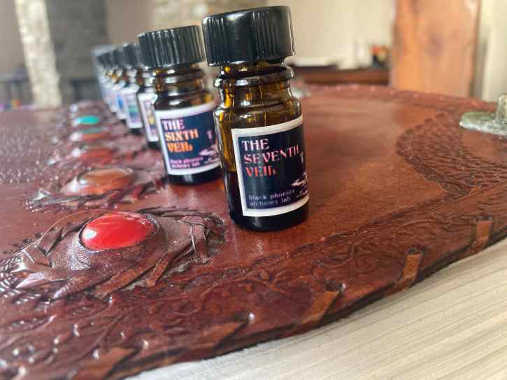 7 5ml perfume bottles arranged next to 7 "chakra" stones, which are set into a big, leather-bound book; the closest perfume bottle, which is the only one in focus, reads "the seventh veil"
